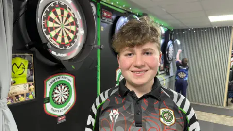 14 year old, Bailey, stands next to a dartboard at Cwm Taf Arrows Darts Academy