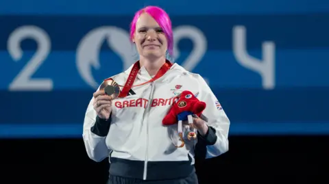 PA Media Great Britain's Jodie Grinham on the podium with the bronze medal after the Women's Individual Compound Open Bronze Medal event at Invalides during day three of the Paris 2024 Summer Paralympic Games. She has pink hair and is holding her medal in her right hand.