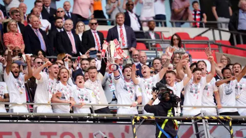 PA Media Crawley Town's Dion Conroy lifts the trophy after the Sky Bet League Two play-off final at Wembley Stadium in London. All the players are wearing white tops and medals.