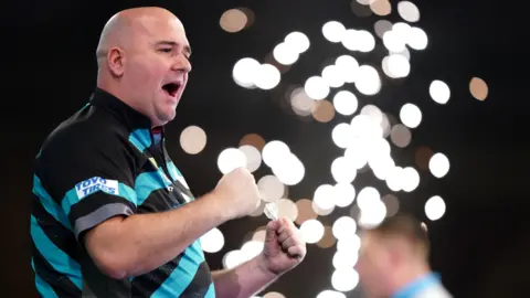 PA Media Rob Cross celebrates after beating Chris Dobey on day fourteen of the Paddy Power World Darts Championship at Alexandra Palace, London. He is wearing a blue and black shirt and is clenching his fists.