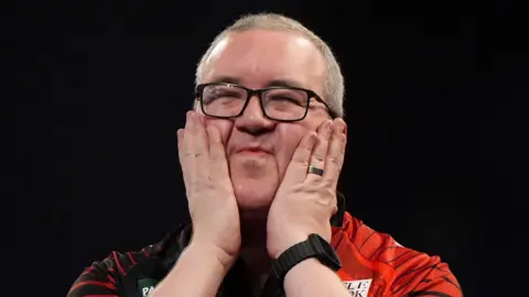 PA Media A man with cropped, greying, fair hair wearing black-rimmed glasses and a red and black darts top. His eyes are closed and he is smiling as he holds his hands up to his face.