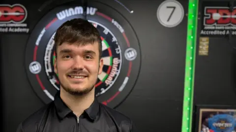 Bailey McNamara smiles as he stands next to a dartboard