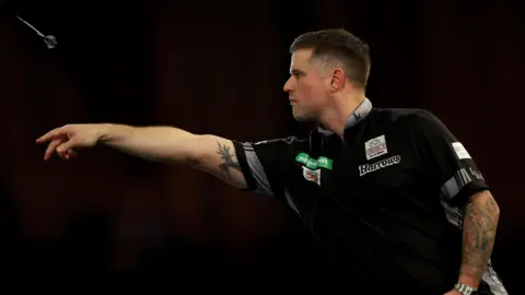 Getty Images A darts player with cropped brown hair and tattooed arms wears a black top with sponsor logos on it. The background is black. His arm is outstretched having just launched a dart which is visible in the top right of the shot.