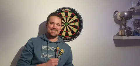 Ian Driver Ian Driver is holding a set of darts and is standing in front of a board and a shelf holding his trophies