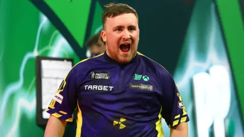 Reuters Luke Littler has his mouth open wide in a smile as he looks off from camera on the darting stage