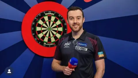 BBC Huw Ware smiles looking at the camera. He is standing in front of a dart board and is holding a mic.