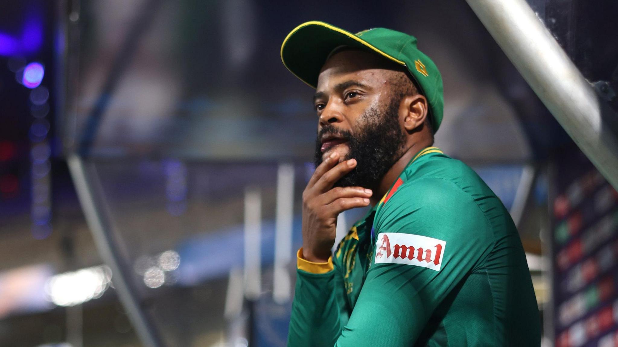 Temba Bavuma, wearing a green South Africa top and baseball cap, looks pensive as he sits with his right hand on his chin in a dugout at the side of a cricket field