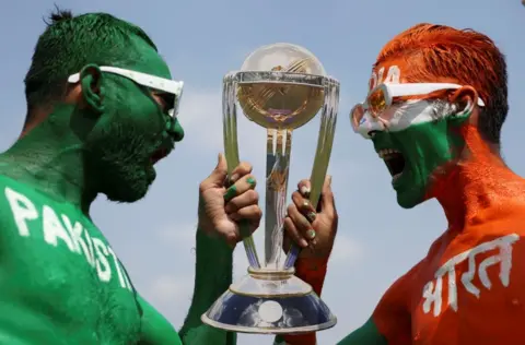 Reuters Cricket fans, Arun Haryani (Right) and Anil Advani (Left) pose for a photograph with a replica trophy after painting their bodies in the Indian and Pakistani national flag colours, ahead of the match between India and Pakistan in the ICC World Cup, in Ahmedabad, India, October 11, 2023.