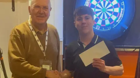 Family handout A man is shaking hands with a boy, who is holding a cheque. Behind them is a darts board.