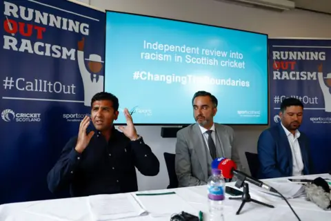 Getty Images Majid Haq and Qasim Sheikh hold a news conference with lawyer Aamer Anwar at the Stirling Court hotel on July 25, 2022 in Stirling. They are dressed casually in dark blue and white open necked shirts and Mr Anwar is in the middle in a grey three-piece suit. There is a mic on the table and Running Out Racism branded boards behind them.