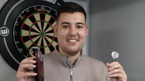 Shaun Whitmore/BBC Ryan Meikle smiles at the camera while standing in front of a dartboard. He is holding an electric hair clipper in one hair and a dart in his other. He has short dark hair and is wearing a grey coloured jumper.