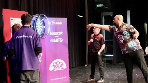 Nescot Professional darts player Devon Petersen throws a dart at a board in front of pupils from Nescot who have their backs turned to camera