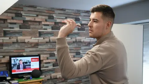 Shaun Whitmore/BBC Meikle is pictured aiming a dart at a dartboard. A TV can be seen behind him with BBC News on.