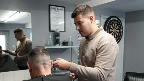 Shaun Whitmore/BBC Meikle is pictured giving a gentleman in a chair a haircut. He is holding a pair of clippers to the man's head.