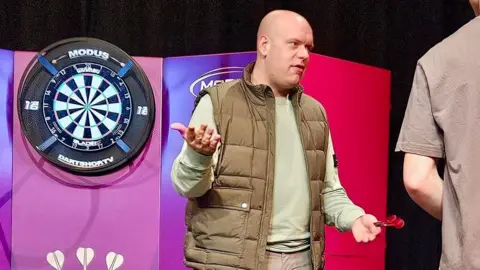 Nescot Michael Van Gerwen standing next to a dart board with darts in his hand at Nescot in Epsom