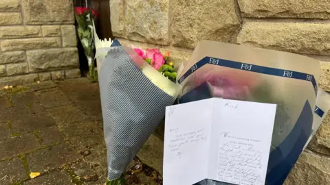 Three floral bouquets are propped up vertically outside the stone wall outside Hatton's home. One of the tem has a condolence card with messages written inside.