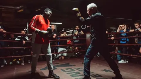 Paul Gilfeather Frank in a boxing ring wearing a dark tracksuit, he is also wearing brown boxing gloves with golden crowns on them. Across from him is one of his students wearing grey joggers and a red top. He appears to be showing him boxing techniques and behind the ropes of the ring there are onlooking students watching the demonstration.